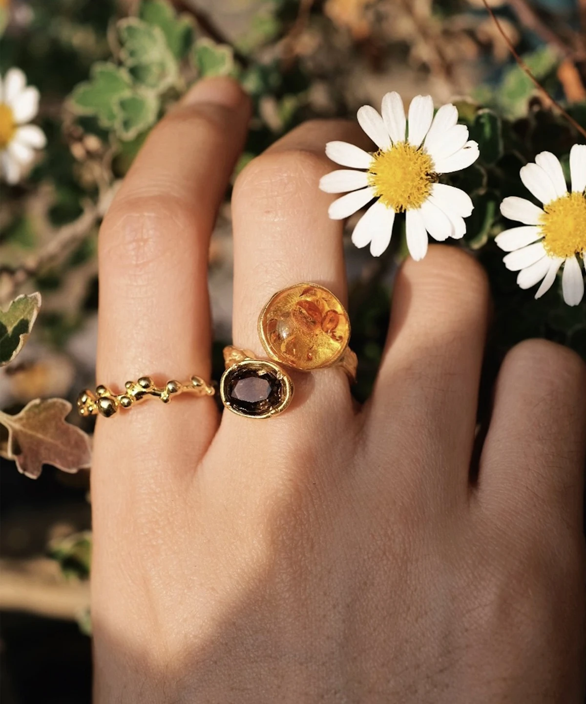 Hand wearing three gold rings, including an Amber Ring with warm golden and deep purple gemstones, shown among white daisies and green foliage.