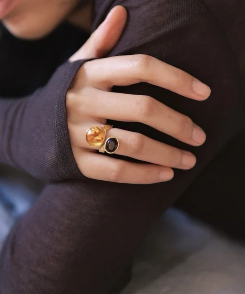 Hand wearing a gold toned Amber Ring with a large amber stone and a smaller dark gemstone, shown against a dark sleeve.