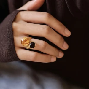 Gold Plated S925 Sterling Silver Amber Ring with Dual Gemstones 06 Hand wearing two gold toned rings, including an Amber Ring with a large amber gemstone and a smaller dark stone, shown against a soft blue‑gray background.