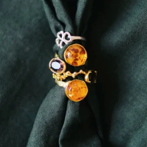 Gold Plated S925 Sterling Silver Amber Ring with Dual Gemstones 09 Five rings displayed on dark green fabric, including multiple gold toned Amber Rings with round amber stones and dark gemstones, arranged beside a silver clover‑shaped ring.