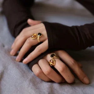 Gold Plated S925 Sterling Silver Amber Ring with Dual Gemstones 11 Two hands wearing matching gold toned Amber Rings with dual gemstones—one amber‑colored and one dark—resting on soft fabric.