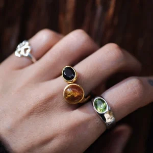 Gold Plated S925 Sterling Silver Amber Ring with Dual Gemstones 14 Hand wearing multiple rings, including a gold toned Amber Ring with a warm amber stone and a dark gemstone, shown alongside silver and green‑stone rings.