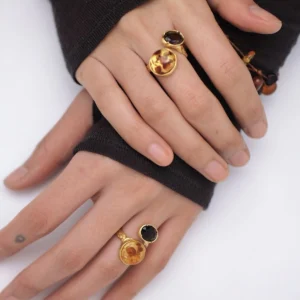 Gold Plated S925 Sterling Silver Amber Ring with Dual Gemstones 17 Two hands wearing gold toned Amber Rings with dual gemstones—a warm amber stone and a dark gem—interlaced against a soft fabric background.