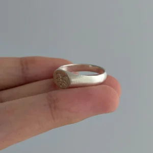 999 Silver Signet Ring with engraved floral or sunburst motif on circular face; handcrafted Silver Signet Ring held between fingers, styled against light gray background to highlight artisanal detail and organic elegance.