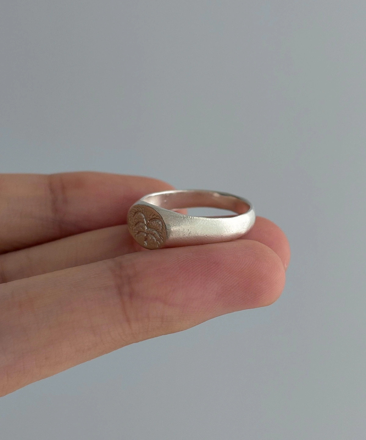 999 Silver Signet Ring with engraved floral or sunburst motif on circular face; handcrafted Silver Signet Ring held between fingers, styled against light gray background to highlight artisanal detail and organic elegance.