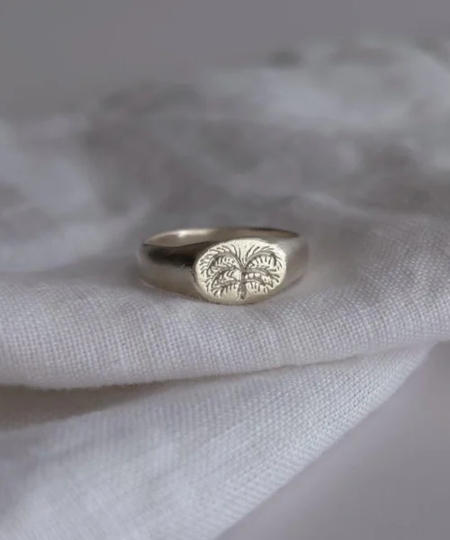 999 Silver Signet Ring with engraved palm tree design on oval face; handcrafted Silver Signet Ring placed on white textured fabric, showcasing tropical symbolism and artisanal detail in close-up view.