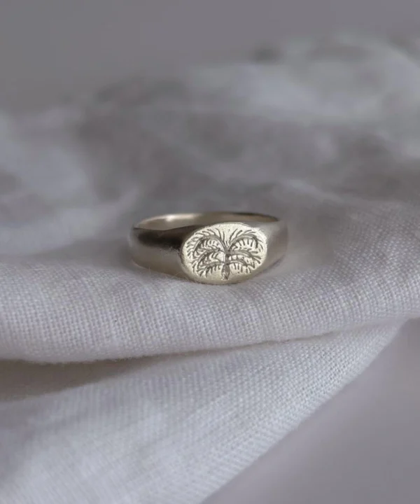 New In 39 999 Silver Signet Ring with engraved palm tree design on oval face; handcrafted Silver Signet Ring placed on white textured fabric, showcasing tropical symbolism and artisanal detail in close-up view.
