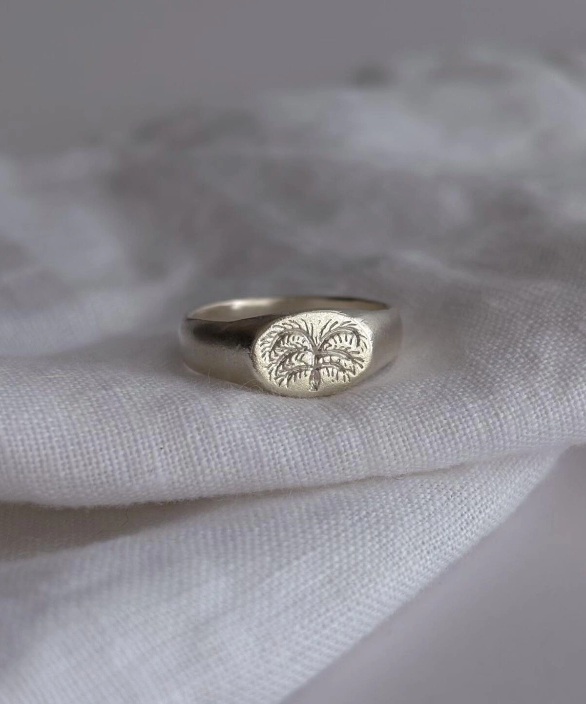 999 Silver Signet Ring with engraved palm tree design on oval face; handcrafted Silver Signet Ring placed on white textured fabric, showcasing tropical symbolism and artisanal detail in close-up view.