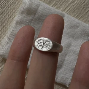 999 Silver Signet Ring with engraved palm tree design on oval face; handcrafted Silver Signet Ring worn on finger, styled against beige fabric and frayed white cloth for a natural, rustic aesthetic.