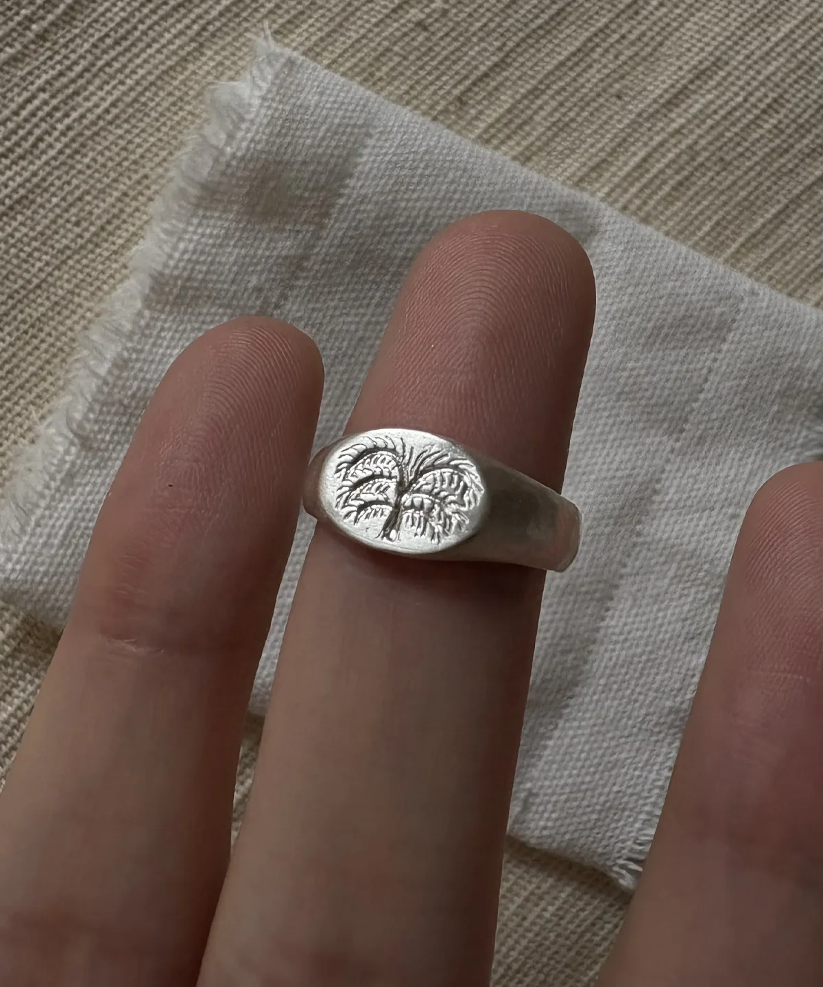 999 Silver Signet Ring with engraved palm tree design on oval face; handcrafted Silver Signet Ring worn on finger, styled against beige fabric and frayed white cloth for a natural, rustic aesthetic.