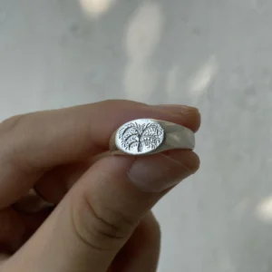 999 Silver Signet Ring with engraved palm tree design on oval face; handcrafted Silver Signet Ring held between fingers, styled against soft neutral background to highlight tropical symbolism and artisanal detail.