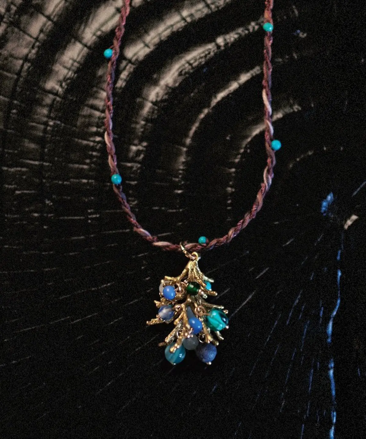 Juniper Berry-Turquoise Pinecone Necklace with braided brown and purple cord, turquoise beads, and gold-toned pinecone pendant; styled on textured dark surface with concentric rings to highlight forest-inspired elegance.
