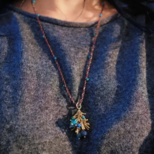 Juniper Berry-Turquoise Pinecone Necklace with braided brown cord, turquoise beads, and gold-toned pinecone pendant; worn over dark textured shirt to highlight forest-inspired elegance and handcrafted detail.