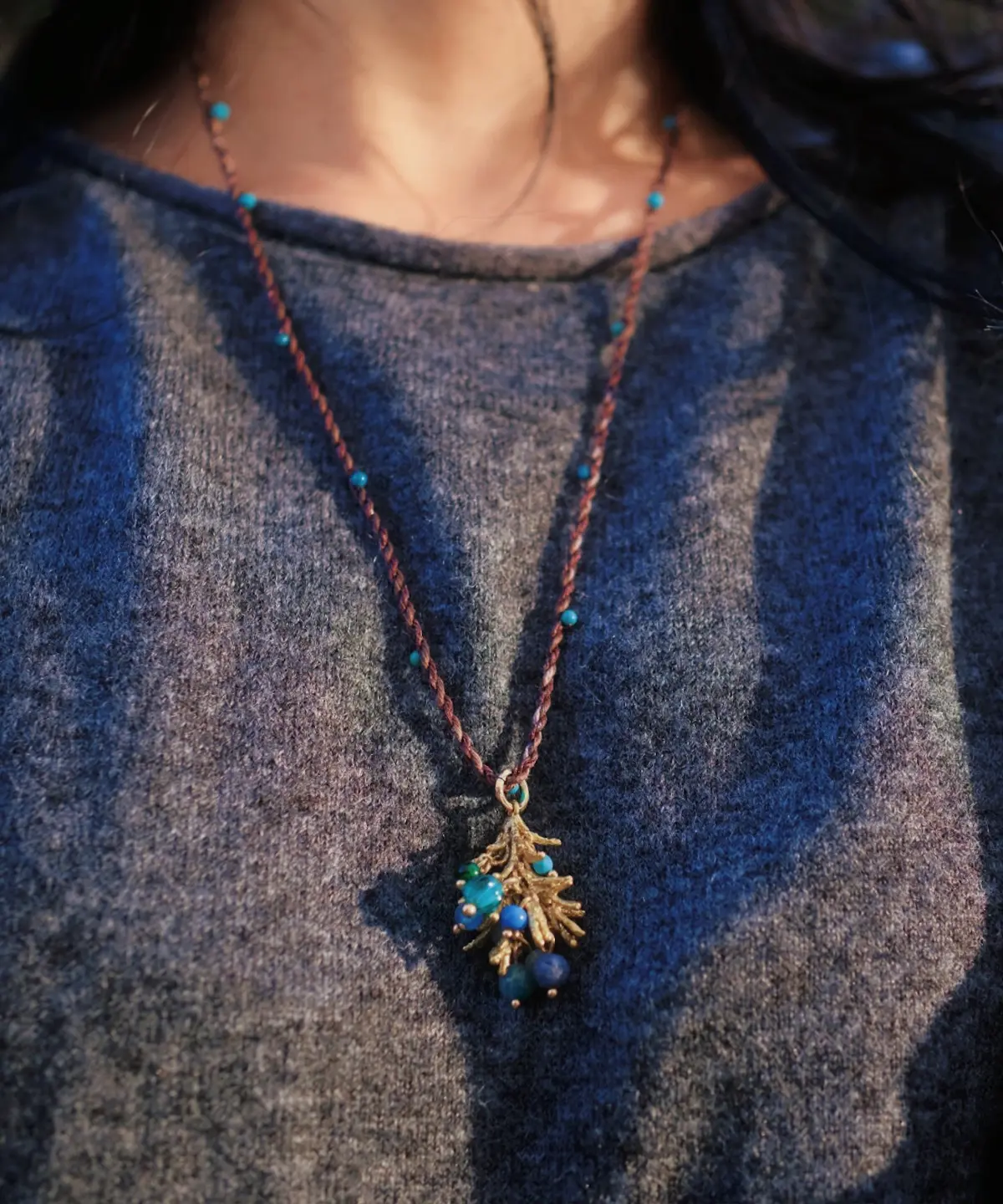 Juniper Berry-Turquoise Pinecone Necklace with braided brown cord, turquoise beads, and gold-toned pinecone pendant; worn over dark textured shirt to highlight forest-inspired elegance and handcrafted detail.