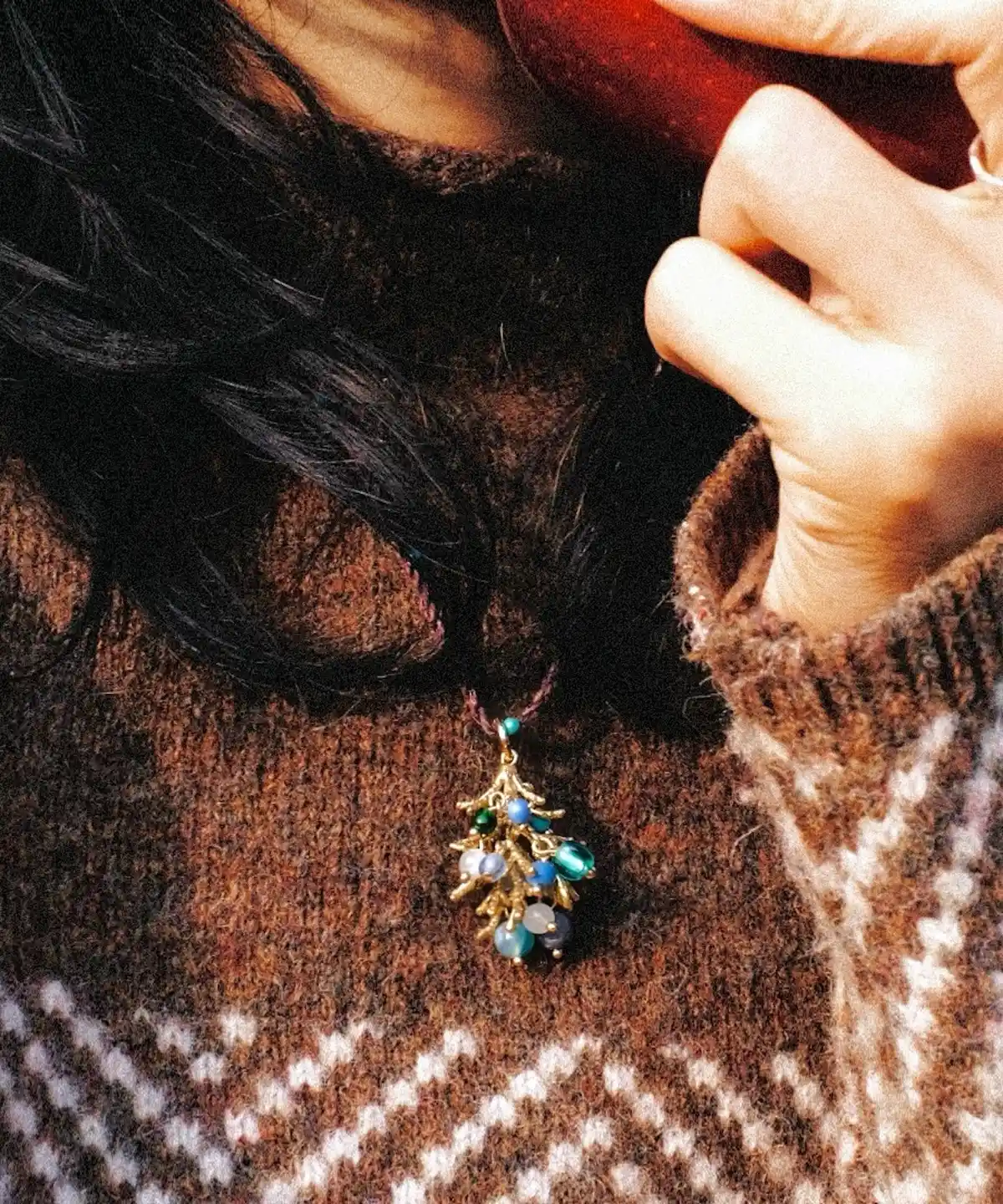 Juniper Berry-Turquoise Pinecone Necklace with braided cord and botanical pendant featuring blue and green beads; worn over brown sweater with white zigzag pattern, highlighting nature-inspired elegance and handcrafted detail.