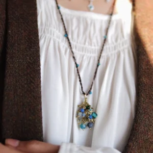 Juniper Berry-Turquoise Pinecone Necklace with braided brown cord, turquoise beads, and gold-toned pinecone pendant; layered with beaded chain over white gathered blouse and brown outerwear, highlighting nature-inspired elegance and handcrafted detail.