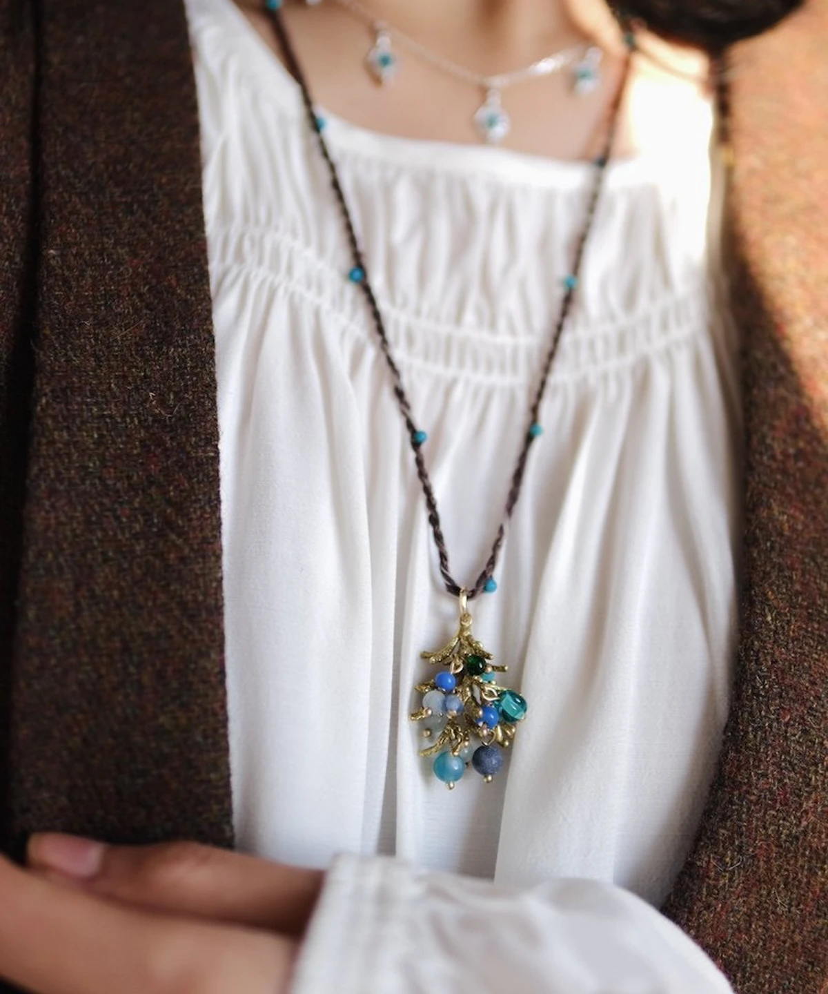 Juniper Berry-Turquoise Pinecone Necklace with braided brown cord, turquoise beads, and gold-toned pinecone pendant; layered with beaded chain over white gathered blouse and brown outerwear, highlighting nature-inspired elegance and handcrafted detail.
