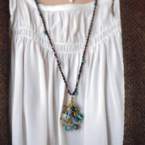 Juniper Berry-Turquoise Pinecone Necklace with braided dark cord, turquoise beads, and gold-toned botanical pendant; layered with silver-toned necklace over white blouse and brown outerwear, highlighting handcrafted woodland elegance.