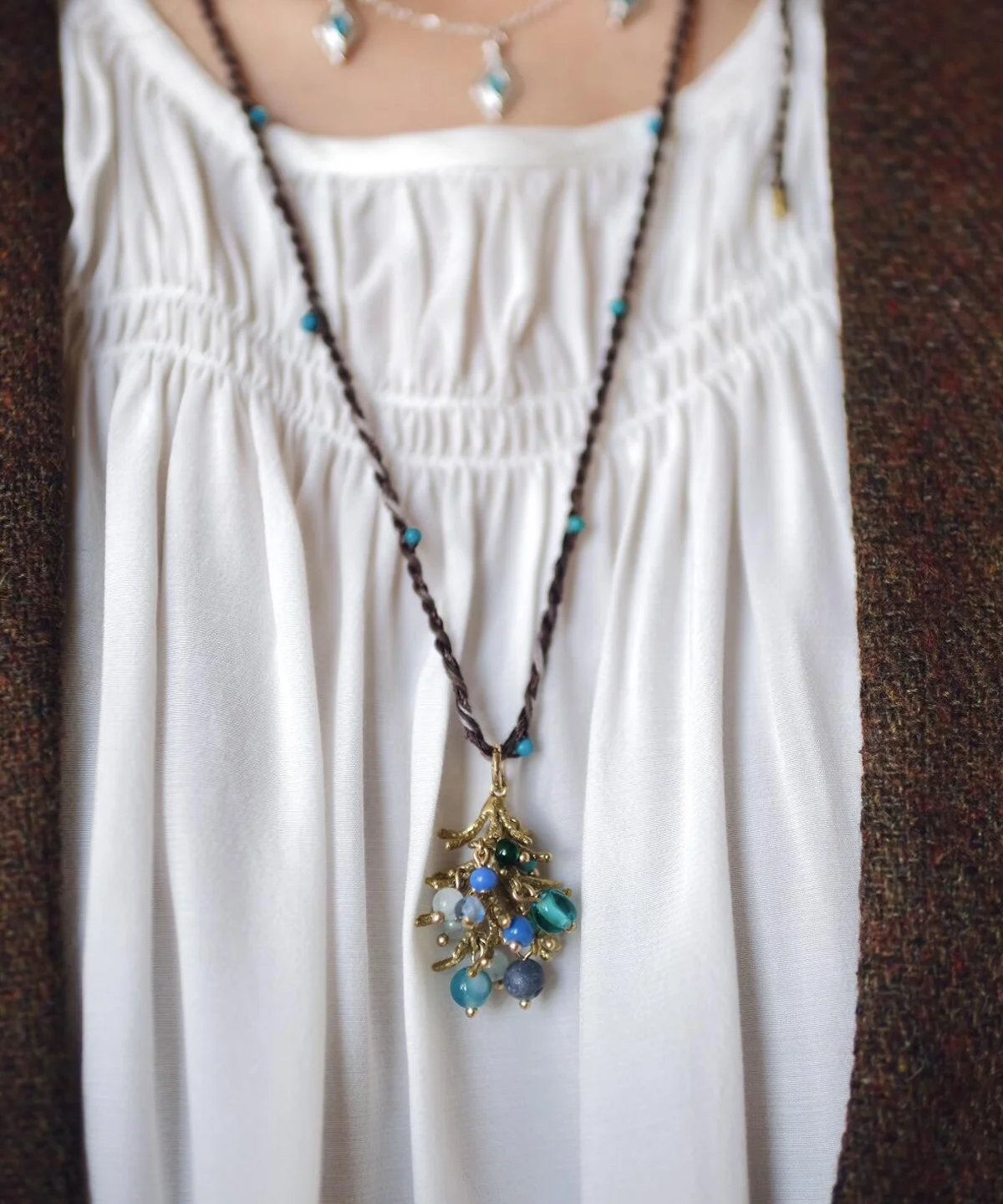 Juniper Berry-Turquoise Pinecone Necklace with braided dark cord, turquoise beads, and gold-toned botanical pendant; layered with silver-toned necklace over white blouse and brown outerwear, highlighting handcrafted woodland elegance.