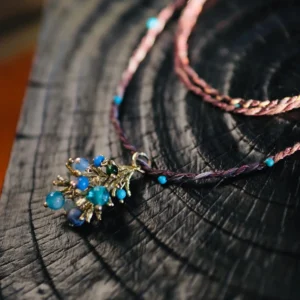 Juniper Berry-Turquoise Pinecone Necklace with purple-pink braided cord, turquoise beads, and gold-toned pinecone pendant; styled on dark wooden surface to highlight handcrafted detail and nature-inspired elegance.
