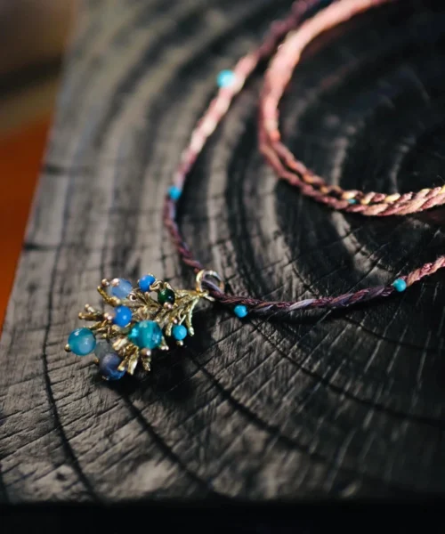 Juniper Berry-Turquoise Pinecone Necklace with purple-pink braided cord, turquoise beads, and gold-toned pinecone pendant; styled on dark wooden surface to highlight handcrafted detail and nature-inspired elegance.