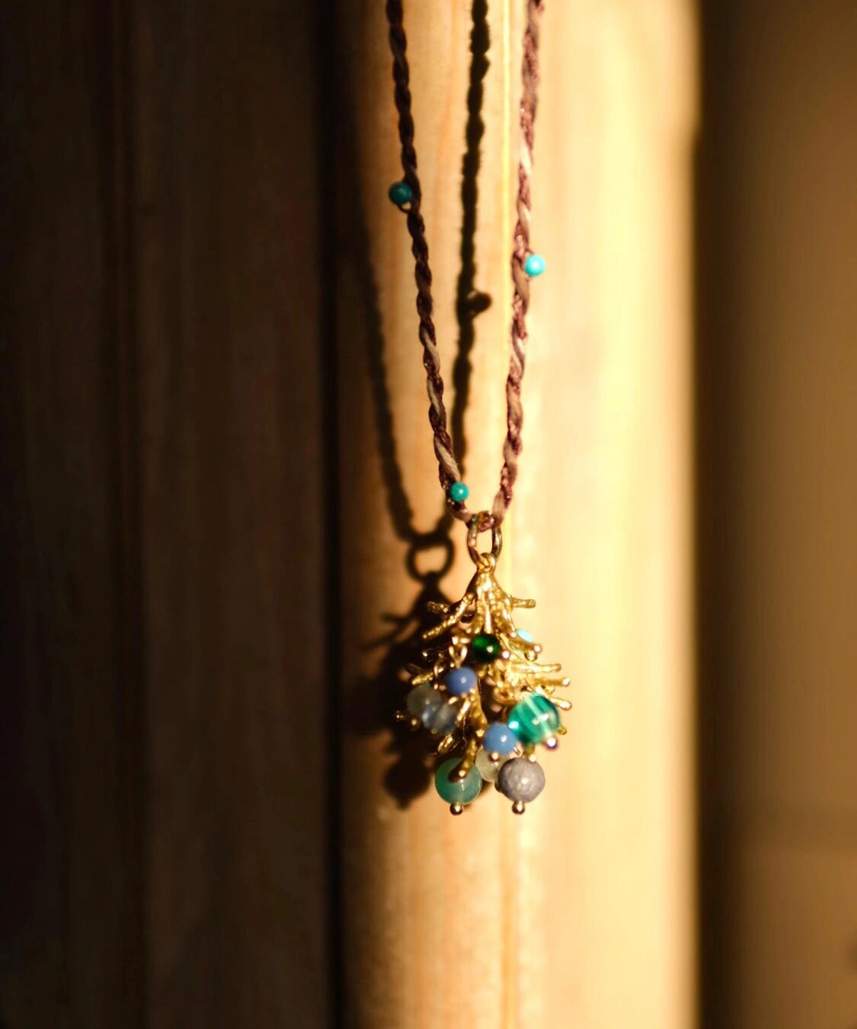 Juniper Berry-Turquoise Pinecone Necklace with twisted brown cord, turquoise beads, and gold-toned pinecone pendant; styled against warm wooden backdrop in sunlight to highlight handcrafted detail and forest-inspired elegance.