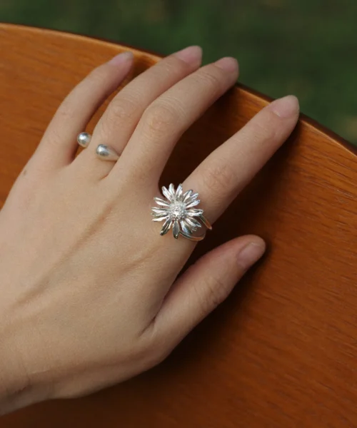 A silver daisy ring with sculpted petals and a textured center, worn on the hand outdoors in soft light.