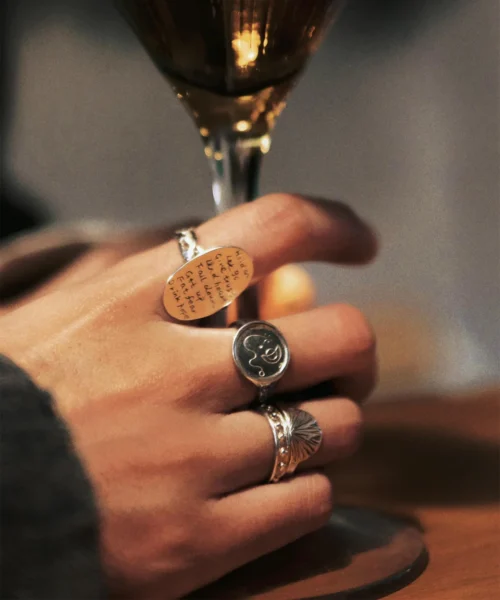 Gold plated Verse Ring with engraved poetic lines, worn on hand holding a golden-stemmed glass; artistic silver rings complement the look.