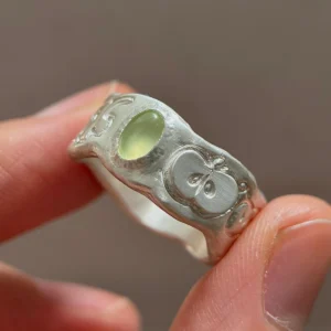 Natural Prehnite Ring in 999 Silver with green oval gemstone and engraved apple motif; a handcrafted 999 Silver Ring featuring symbolic leaf and seed details, held between fingers in close-up view.