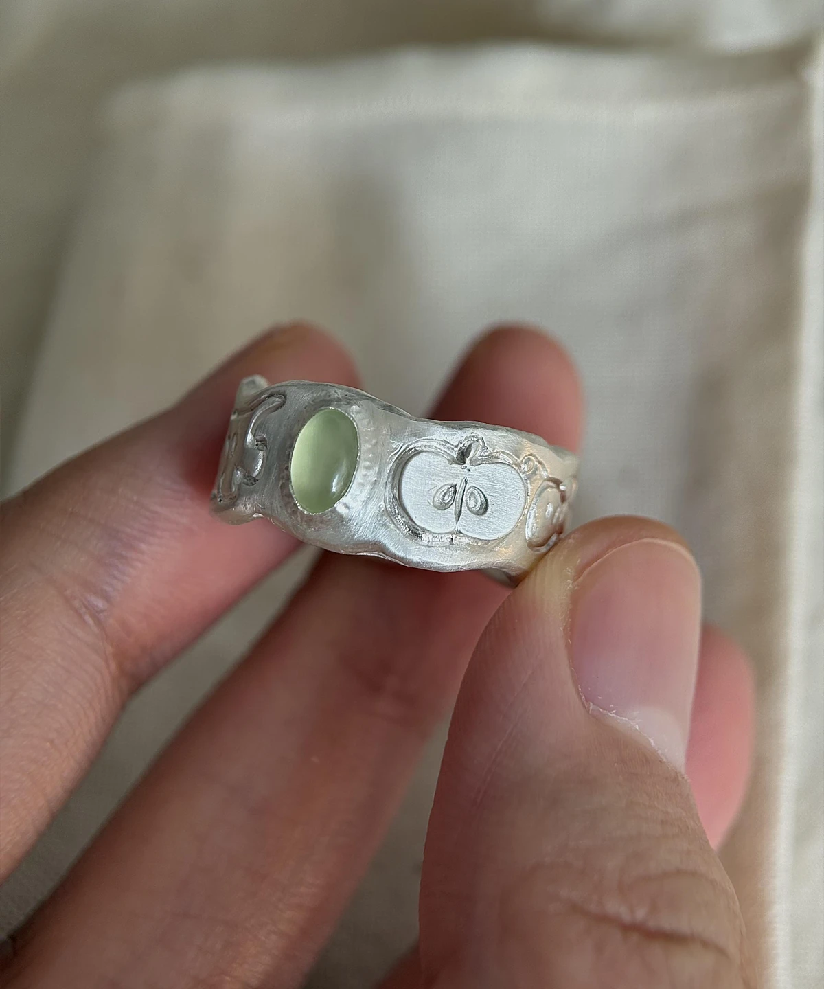 Natural Prehnite Ring in 999 Silver Green Apple Design 02 Natural Prehnite Ring in 999 Silver featuring green oval gemstone and engraved apple motif with leaf and seeds; a handcrafted 999 Silver Ring held between fingers in close-up view, showcasing symbolic and organic design.