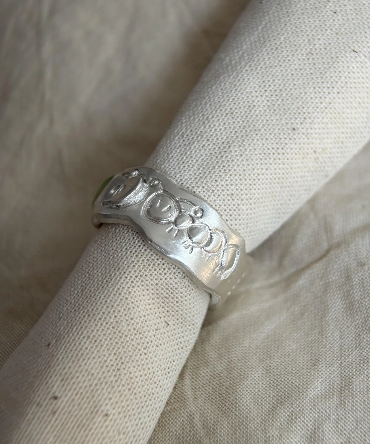 Natural Prehnite Ring in 999 Silver Green Apple Design 09 A handcrafted 999 silver band engraved with a row of whimsical stick‑figure faces, photographed on soft beige fabric. A playful artisan piece paired with the Natural Prehnite Ring style.