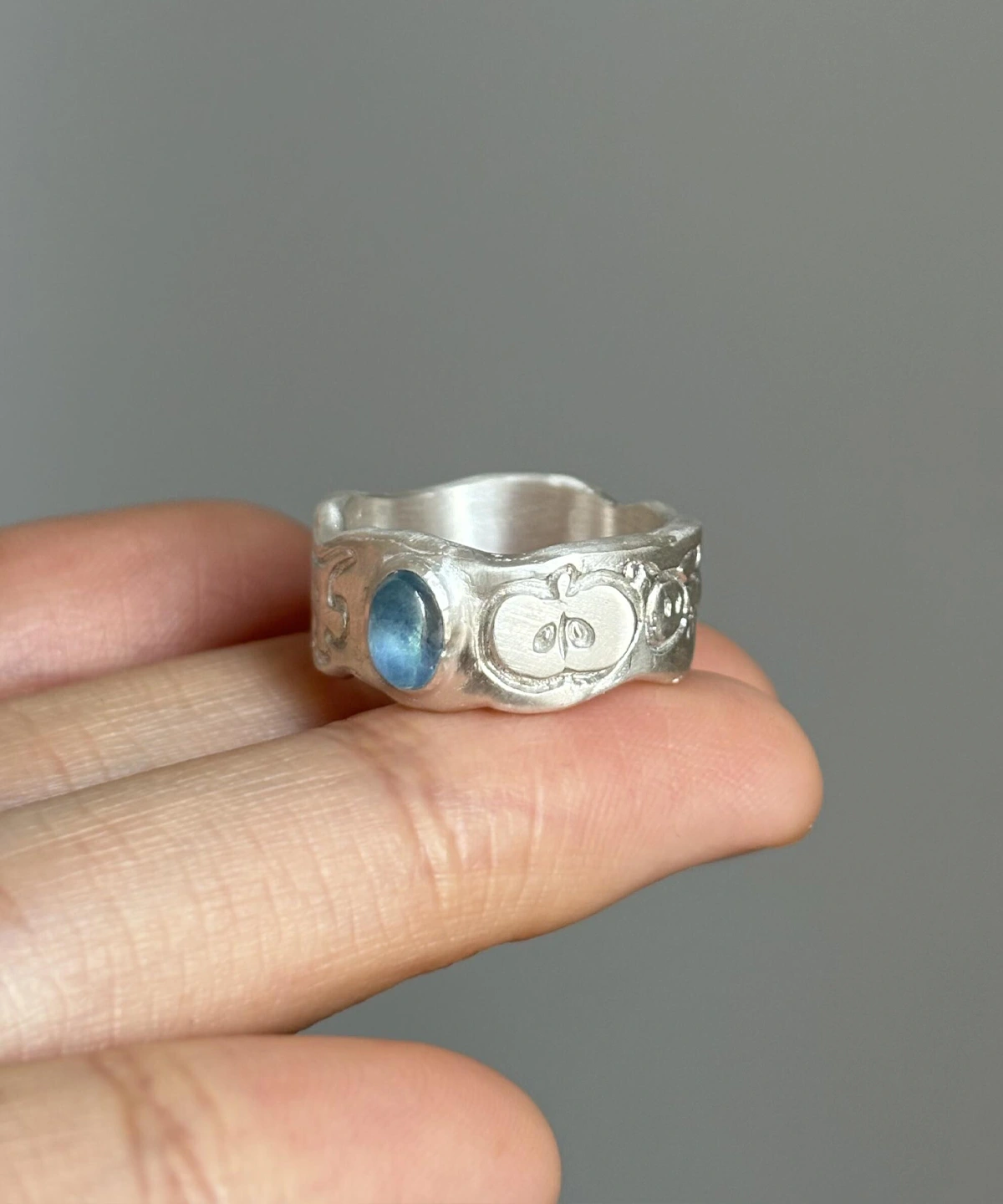 Natural Prehnite Ring in 999 Silver Green Apple Design 13 A handcrafted 999 silver ring featuring a blue oval aquamarine gemstone and engraved apple‑inspired symbols, held in hand to highlight the artisanal Natural Aquamarine Ring design.