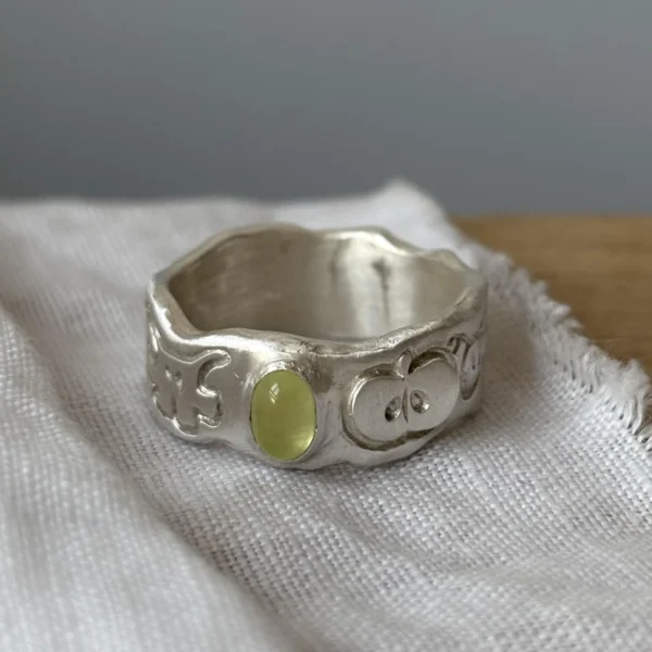 Home 56 A handcrafted 999 silver ring with an organic engraved band and a yellow‑green oval gemstone, displayed on textured white fabric, styled to complement the Natural Prehnite Ring aesthetic.