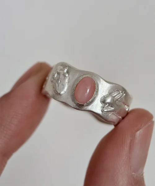 Hand holding a handcrafted silver ring with a pink oval gemstone, featuring detailed ladybug and rabbit motifs on the band.