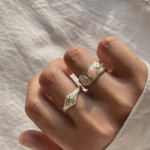 Hand wearing two handcrafted silver rings, including a Pink Opal Ring with an oval pink stone and sculptural metalwork, shown against textured white fabric.