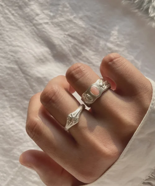 Hand wearing two handcrafted silver rings, including a Pink Opal Ring with an oval pink stone and sculptural metalwork, shown against textured white fabric.