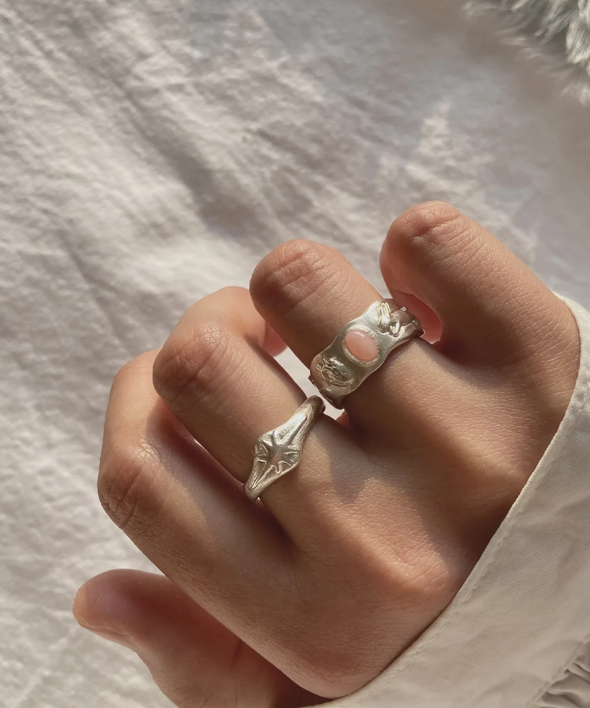 Hand wearing two handcrafted silver rings, including a Pink Opal Ring with an oval pink stone and sculptural metalwork, shown against textured white fabric.