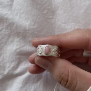 Strawberry Thief-Handmade 999 Silver Pink Opal Ring 05 Hand holding a handcrafted silver Pink Opal Ring with an oval pink stone and detailed engraved motifs on each side, shown against soft white fabric.