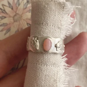 Strawberry Thief-Handmade 999 Silver Pink Opal Ring 09 Hand holding a handcrafted silver Pink Opal Ring with an oval pink stone and embossed strawberry and mushroom motifs, displayed on textured fabric.