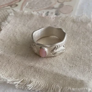 Hand holding a handcrafted silver Pink Opal Ring with an oval pink stone and raised rabbit‑inspired motifs on a wide, wavy band, displayed on textured fabric.