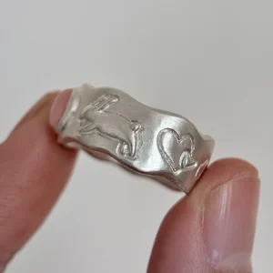 Strawberry Thief-Handmade 999 Silver Pink Opal Ring 17 Hand holding a handcrafted silver ring with engraved rabbit and heart motifs on a wavy band, shown without the central Pink Opal Ring stone.