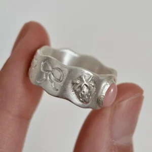 Hand holding a handcrafted silver Pink Opal Ring with a round pink stone and engraved bow and anatomical‑heart motifs on a wavy textured band.
