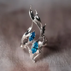Sterling Silver Statement Ring with twin marquise-cut blue gemstones and flowing fishtail-inspired silver curves; handcrafted ring placed on neutral surface, showcasing dynamic design and oceanic elegance.