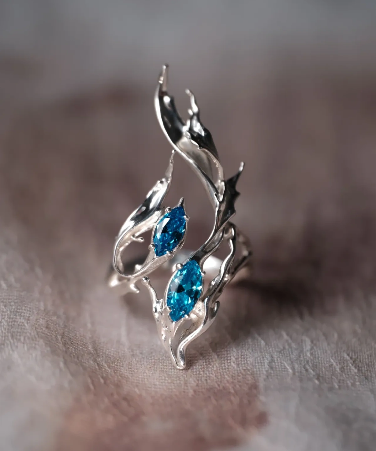 Sterling Silver Statement Ring with twin marquise-cut blue gemstones and flowing fishtail-inspired silver curves; handcrafted ring placed on neutral surface, showcasing dynamic design and oceanic elegance.
