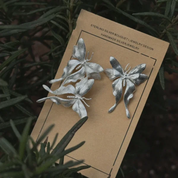 Asymmetric Butterfly Earrings in S990 Sterling Silver 02 Handcrafted asymmetric Butterfly Earrings in S990 sterling silver displayed on a beige card with surrounding green foliage, featuring sculpted floral textures and artisanal details.