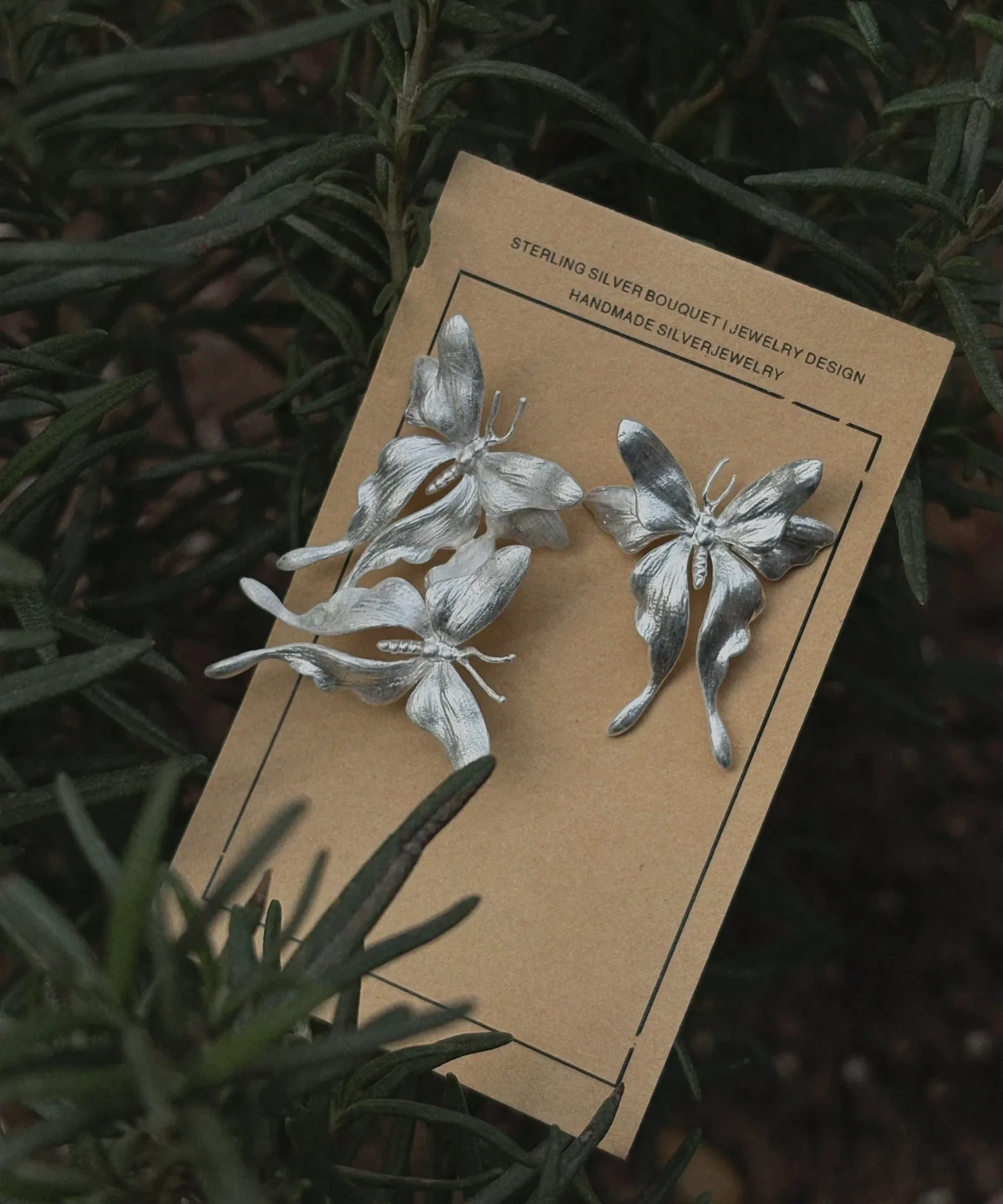 Asymmetric Butterfly Earrings in S990 Sterling Silver 02 Handcrafted asymmetric Butterfly Earrings in S990 sterling silver displayed on a beige card with surrounding green foliage, featuring sculpted floral textures and artisanal details.