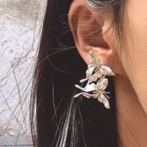 Asymmetric Butterfly Earrings in S990 sterling silver worn on the ear, featuring sculptural multi-flower details and textured petals framed by dark hair in a softly blurred outdoor setting.
