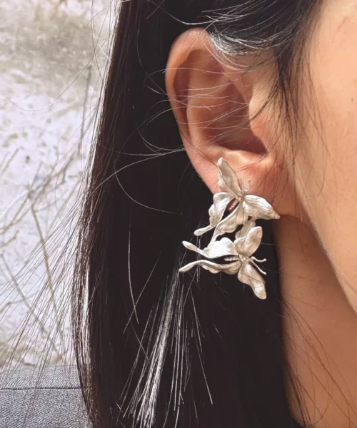 Asymmetric Butterfly Earrings in S990 sterling silver worn on the ear, featuring sculptural multi-flower details and textured petals framed by dark hair in a softly blurred outdoor setting.