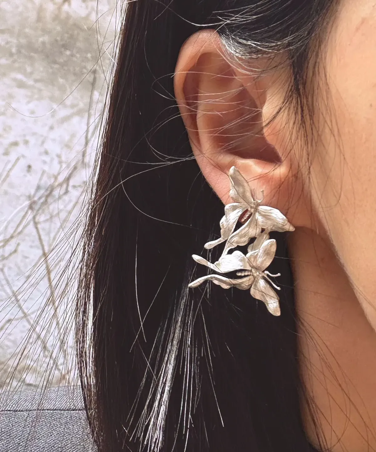 Asymmetric Butterfly Earrings in S990 sterling silver worn on the ear, featuring sculptural multi-flower details and textured petals framed by dark hair in a softly blurred outdoor setting.