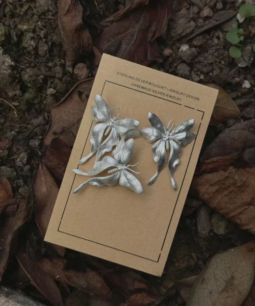 Three handcrafted Butterfly Earrings in S990 sterling silver displayed on a card placed among dried leaves and soil, featuring sculpted wings and detailed textures in a nature-inspired setting.